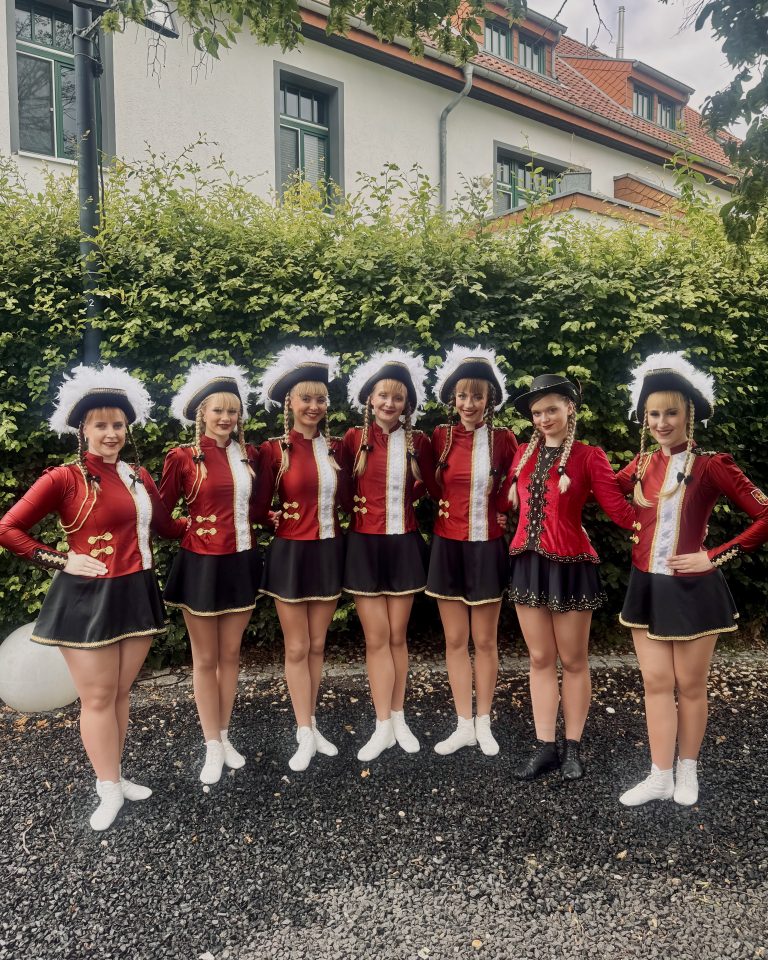 Auf dem Bild sieht man 7 Personen. Sie stehen nebeneinander vor einer grünen Hecke. Sie tragen rote Gardeuniformen und lachen in die Kamera.